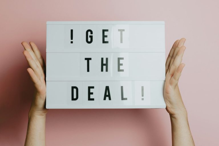 Hände halten ein Schild mit der Aufschrift „GET THE DEAL!“ vor einem rosa Hintergrund.