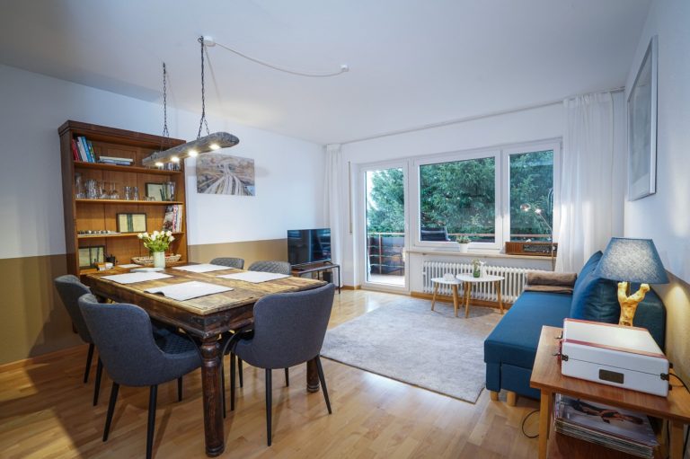 Helles Wohnzimmer mit Esstisch, Stühlen, Bücherregal und einer Couch. Großer Fensterbereich.