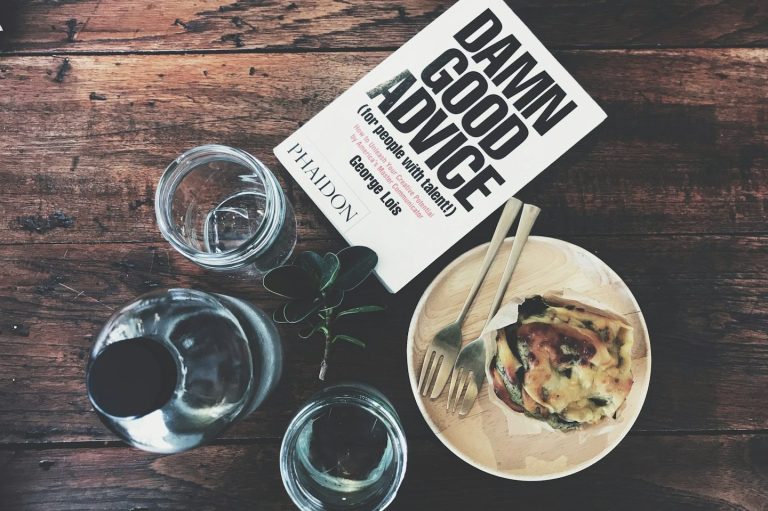 Ein Buch mit dem Titel "Damn Good Advice" liegt neben einem Teller und Gläsern auf einem Holztisch.